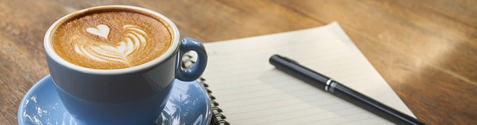 Auf einem Holztisch liegt ein Notizblock mit einem Stift und links steht eine blaue Kaffeetasse.