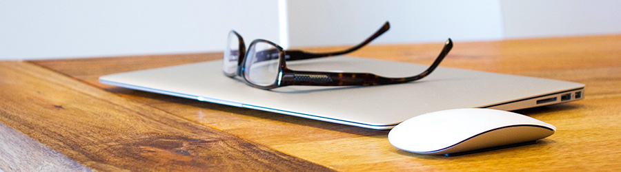 Auf einem Holztisch liegen ein Apple Laptop sowie eine Apple Maus und eine Brille.