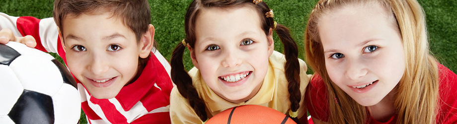 Drei junge lächelnde Kinder gucken in die Kamera und halten ein Fußall und einen Basketball in den Händen.
